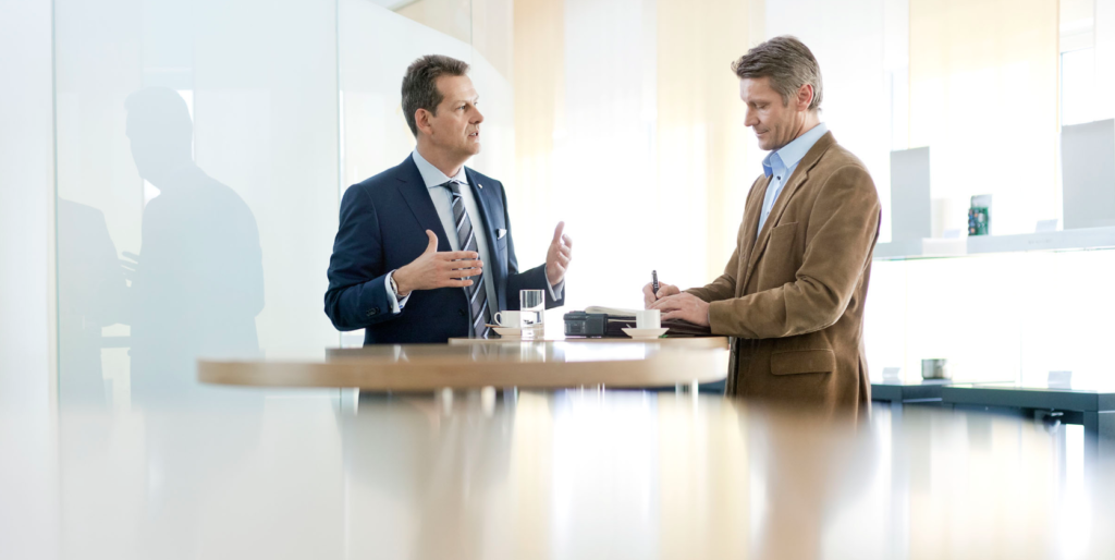 Zwei Männer im Gespräch an einem Stehtisch, einer spricht gestikulierend, der andere macht Notizen